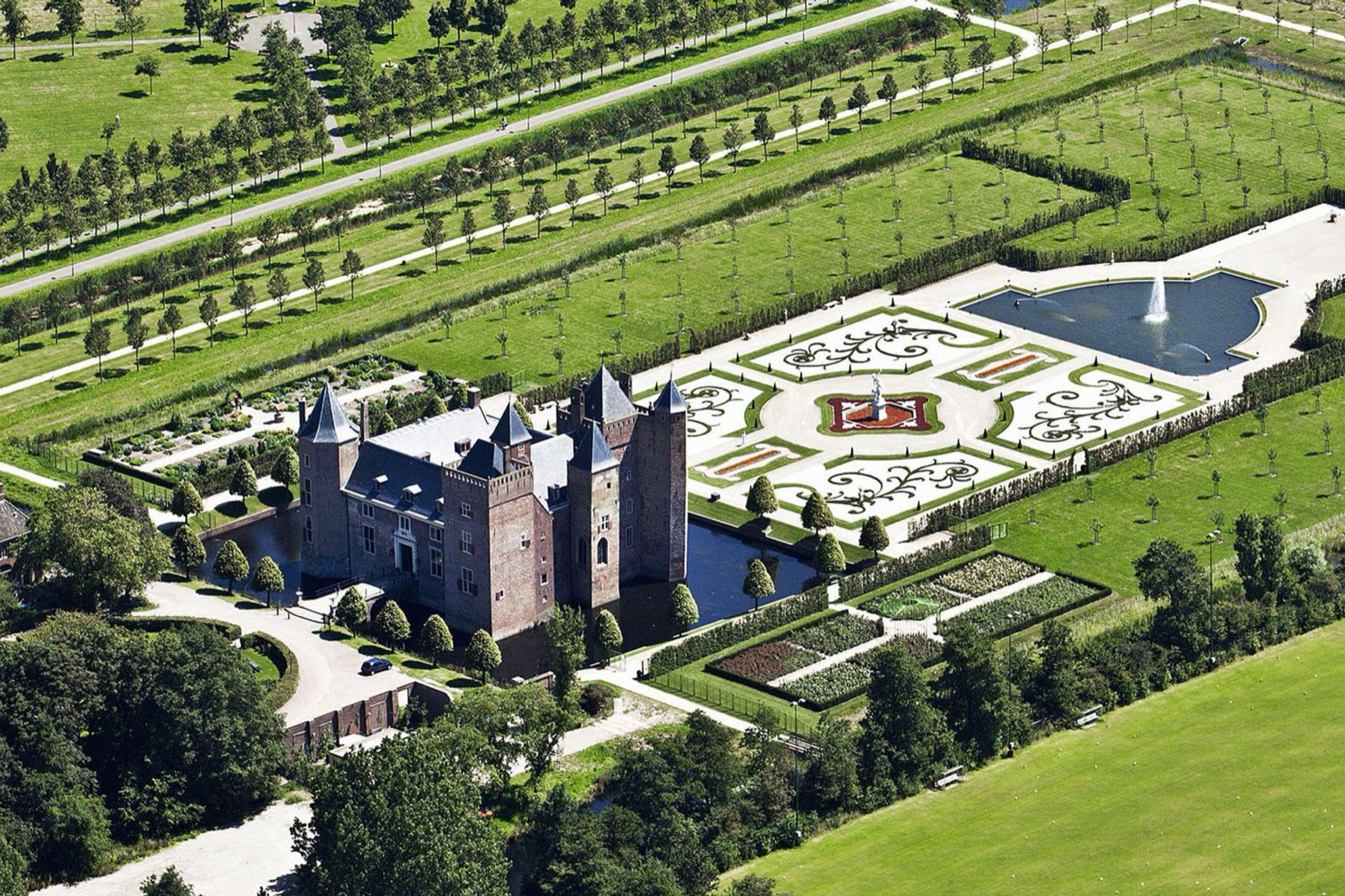 Slot Assumburg van bovenaf: het omgrachte kasteel met daarnaast de formele tuin met fontein, omringd door parkbomen en weilanden.